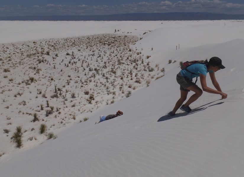 Norma sliding down on her belly while Carmen climbs the dune