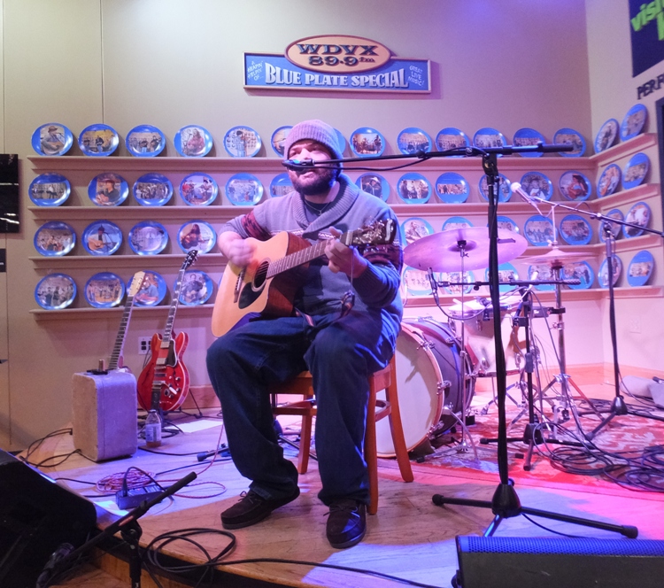 Scruffy man seated, playing guitar and singing